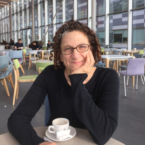 Cynthia Dwork sits at a cafe table in front of a finished cup of coffee. She has fair skin and curly auburn hair with hints of grey. She wears glasses and is smiling.